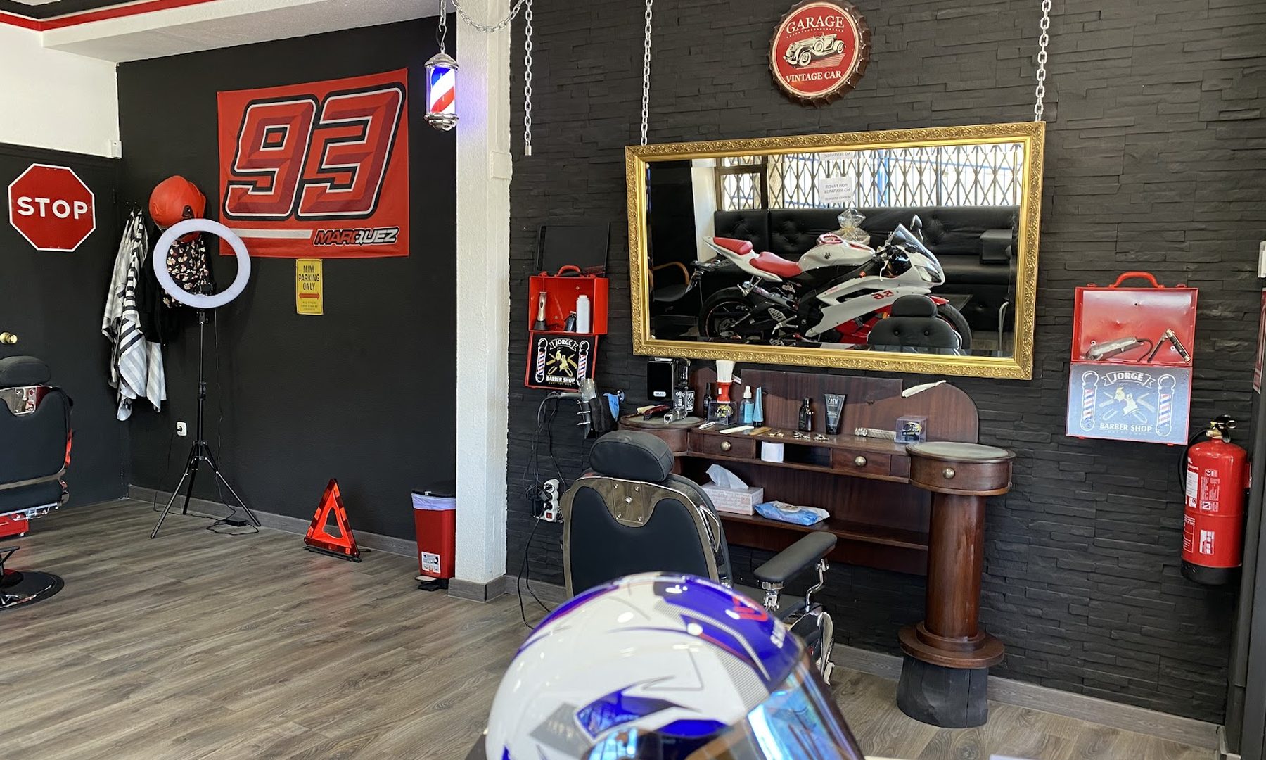 Interior de Jorge Barber Shop con una moto deportiva blanca y roja aparcada entre los sillones de barbero, la pared negra con las gráficas del 93 y una señal de stop.